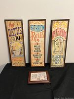Three vertical wooden framed vintage bathroom signs with colorful text and illustrations sitting upright.
