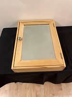 Front view of the wooden medicine cabinet showing mirrored door, metal knob, and light wood finish.