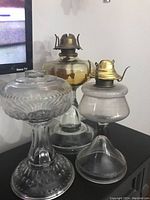 Photo of three vintage oil lamps including an amber-tinted glass lamp with brass burner, and two clear glass oil lamps with detached globe covers, all placed on black surface.