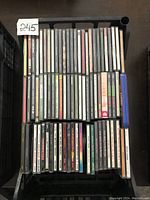 Top-down photo of a black plastic crate filled with approx. 40 music CD jewel cases, stacked vertically in rows. Visible album spines with various artist and album names.