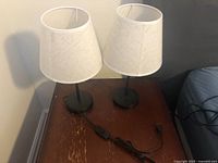 Two small bedside lamps with off-white linen fabric shades and black metal bases sitting on a wooden nightstand. Switches are inline on cords.