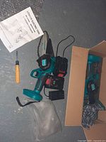 Photo showing a 6 inch handheld lithium electric chainsaw, charger with battery, screwdriver, blade cover, gloves, and user manual all laid out on a concrete floor; chainsaw in teal and black