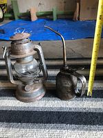 Beacon metal oil lantern and vintage metal oil can displayed on carpet with blue tarp background, showing both items and their scale against a measuring tape.