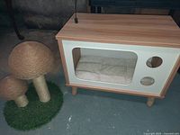 Photo showing a wooden cat house with white panel and clear window, containing a beige cushion. Next to it is a cat scratcher shaped like two mushrooms wrapped in sisal rope on an artificial grass base.