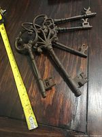 Set of 5 cast iron inspired decorative keys showing various ornate bow designs, placed on wooden surface with measuring tape indicating length approximately 5-7 inches.