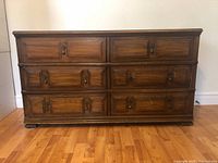 Front view of the wooden dresser showing six drawers with decorative metal handles and keyhole details, standing on a hardwood floor.