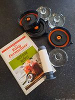 Photo showing two black plastic fermentation lids with orange valve seals, three glass fermentation airlocks, a white and grey fermentation pump, and a user guide booklet titled 'The Easy Fermenter'.