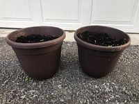 Two identical brown flower pots filled with soil, placed on gravel ground in front of a white garage door.