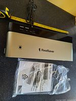 Closed FoodSaver vacuum sealer machine showing silver top with brand logo and buttons, measuring tape placed to indicate approx length.
