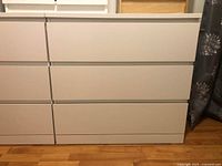 Photo of white Ikea STORKLINTA dresser showing front view with three wide drawers, placed on wood flooring with curtain in background.