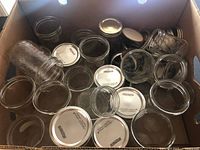 Cardboard box filled with various sizes of clear glass Mason jars, some lying on their sides and others upright, with multiple metal jar lids spread across the box.