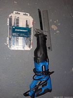 Mastercraft blue and black reciprocating saw with cord, a plastic case containing new blades, and a metal ruler placed together on the floor.