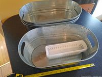 Stacked oval galvanized metal storage baskets with built-in handles shown on a kitchen counter. One basket contains a small white perforated plastic organizer for scale. Yellow tape measure shows dimensions around 17-18 inches long.