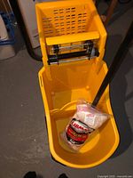 Yellow mop bucket with wringer showing interior and mop handle inside, new mop head package resting inside the bucket