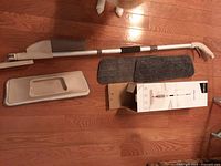 Photo showing Decorus spray mop components including handle with spray trigger, cleaning solution container, two gray microfiber mop pads, and product box.