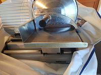Photo showing front view of Nesco slicer, with serrated circular blade, adjustable thickness knob, metal base, sliding food carriage with metal spikes, and clear plastic food guard.