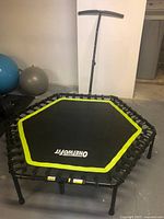 Mini trampoline with hexagonal black jump mat with yellow trim and adjustable black handle bar, shown standing on six legs in a room with exercise balls in the background.