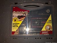 Clear plastic carry case with the Milescraft PocketJig 200 kit visible inside, showing parts and the label indicating it is a complete pocket hole kit with a T20 driver and screw head.