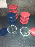 Twelve clear round glass baking dishes with plastic lids in red and blue, stacked on a kitchen countertop. Some lids removed from dishes and placed beside.