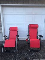Front view of two red reclining lawn chairs with metal frames and attached headrest pillows, placed on gravel in front of garage door.