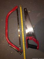 Photo of two hand saws on a grey floor with measuring tape placed to show blade length. Left is a red bow saw, right is a traditional handsaw with a black and red handle.