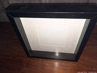 Black square shadow box style picture frame with clear glass front and white interior matting on wood surface.