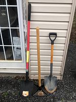 Photo shows two shovels and a steel tamper leaning against the exterior wall of a house by a door. One shovel has a black pointed blade with wooden handle and black D-grip, the other shovel has a red and grey handle and a square blade. The steel tamper has a wooden handle and flat square steel base approximately 10x10 inches.