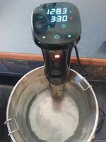 Sous vide immersion cooker attached to pot showing digital temperature of 128.3°F and timer 3:30 on screen.