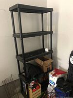 Black plastic four-tier storage shelf placed in a corner with some items around it, shelf empty except for a small round white clock on the second shelf