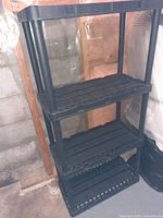 Photo of one black plastic storage shelf with four slatted tiers and cylindrical support legs, located in a basement or garage setting.