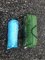 Photo showing two packed items: a blue cylindrical bag labeled 'SUNBABAY' and a green rectangular bag with handles, placed on gravel surface.