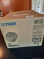 Original box with Tiger branding and model number JPE-A100W visible, sealed box shown on table.