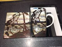 Ceramic mug in box featuring landscape artwork print by Tom Thomson, showing silhouette of tree on shoreline against lake and sky.