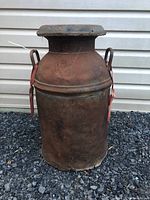 Full front view of vintage milk can on gravel surface against siding wall, shows rust and wear.