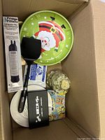 Box with refrigerator water filter, plastic spatula, Santa Claus festive tray, dish with black holder, glass jar with flowers and other small items.