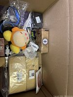 Top-down view of box contents showing orange/yellow plush toy, black pouch, and assorted packaged items