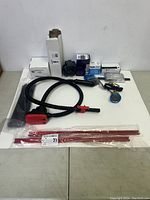 Overview of the miscellaneous lot showing hoses, red metal parts, gauge, and various boxed components arranged on white surface.
