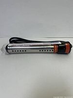 Silver metal cylindrical immersion rod with ventilation holes and attached black power cord, lying horizontally on a white background, showing label and warning instructions.