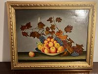 Full view of the framed oil painting showing bowl of fruit and grapevine against muted background.