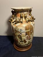 Front view of the ceramic vase showing a painted scene with figures and nature elements, mounted on a wooden base.