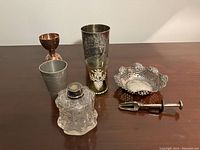 Full view of all items including the sterling nut basket, Victorian olive grabber, copper double jigger, two metal cups, glass cup with painted decoration, and ink bottle.
