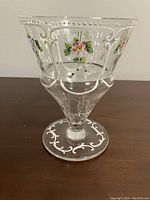 Clear glass goblet with white scroll patterns and hand-painted pink and green flowers on the side and base, photographed from the side.