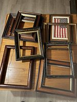 Top-down photo showing a pile of ten medium-sized picture frames arranged overlapping on the floor. One frame contains an American flag with a white mat. The frames vary in color and profile, mostly wooden with some black and gold elements.