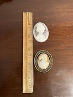 Two carved cameos and a ruler for size reference, showing measurements about 1.5 inches tall each. One cameo is set as a brooch with an ornate frame, the other is loose.