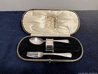 Open case showing sterling silver fork, spoon, and silverplate napkin ring arranged inside on black velvet base and satin-lined lid.