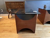 Full front view of teak wood side table with glass top, showing rectangular table with dark semi-circular accents and wood grain visible, placed on hardwood floor near antique wooden furniture.