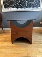 Front angle view of the teak side table with geometric cutouts and glass top, showing wood grain and dark curved accent.