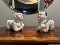 Pair of ceramic baby Buddha figurines placed on wood surface under a round mirror, showing side profile facing each other.