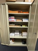 Open metal office storage cabinet with shelves containing orange binders, white and brown boxes, and other office supplies.