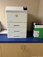 White HP LaserJet Enterprise M507dn printer with three paper trays and digital control panel on a blue countertop next to a box of paper supplies.